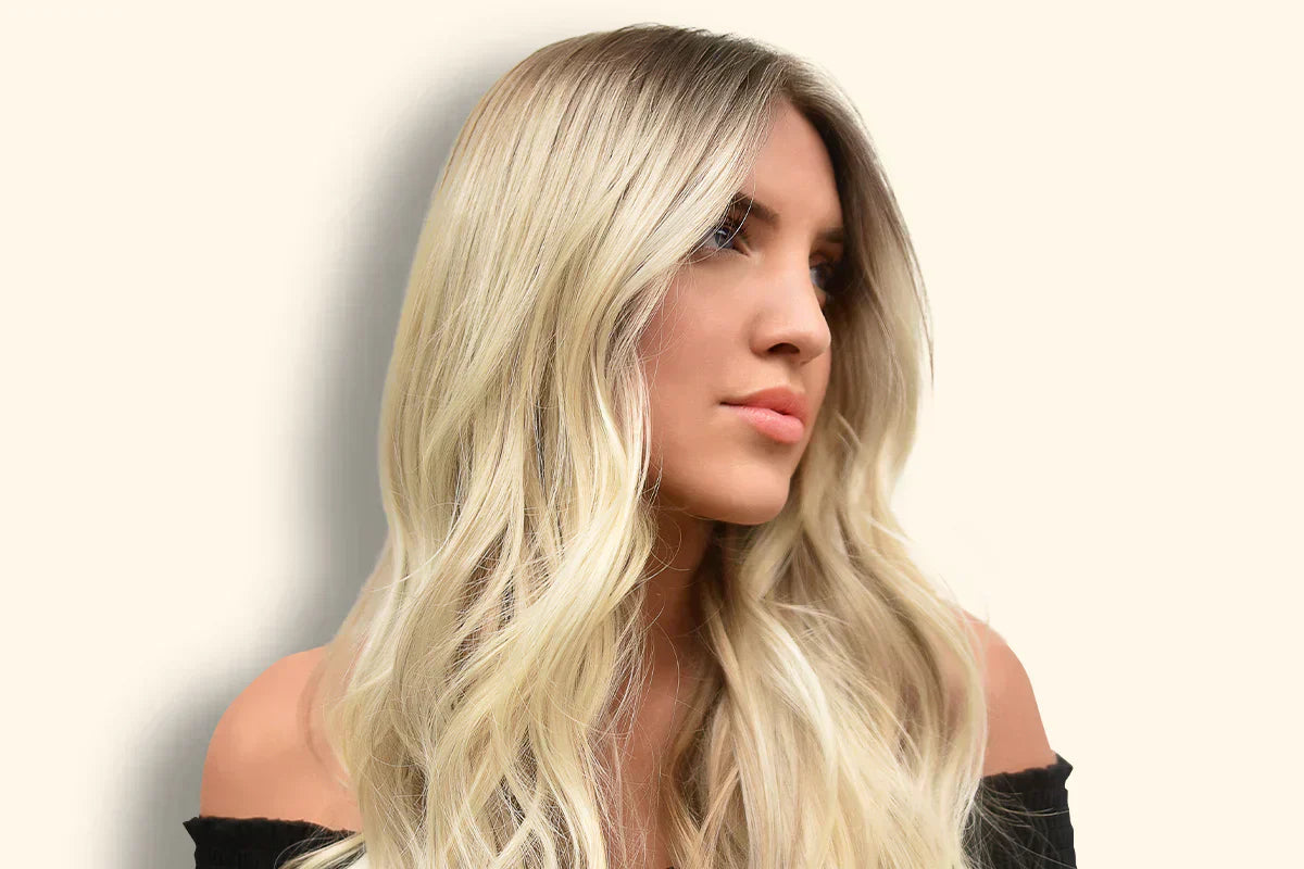 A woman with long, wavy ash blonde hair styled elegantly, wearing an off-the-shoulder black top, against a soft beige background.